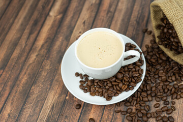 A cup of creamy coffee with roasted coffee beans next to it, wooden background