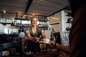 Beautiful young female waiter serving cup of coffee to customer in modern cafe