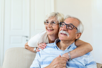 Portrait of overjoyed elderly husband and wife sit on couch hugging looking in distance. Happy mature spouses couple embrace cuddle relax together on sofa, enjoy time together