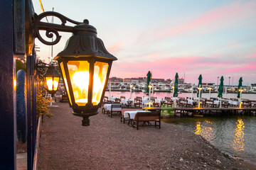 Deserted promenade with piers and cafes during beautiful fiery sunrise or sunset on the sea coast. Elegant street lamp is lit in foreground. © Masarik
