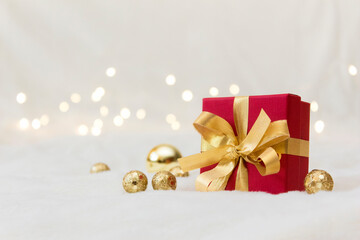 Red gift box with a gold bow on a light background.Yellow Christmas balloons and a garland can be seen in the background.