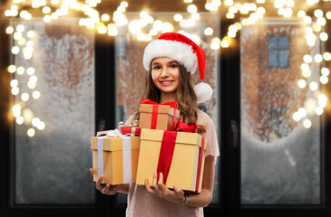 Obraz na płótnie Canvas christmas, holidays and people concept - happy smiling teenage girl in santa helper hat holding gift box over garland lights on snowy window background