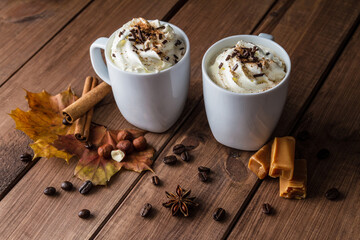Two cups of delicious coffee with whipped cream and chocolate sprinkles. Autumn composition on a wooden table, with cinnamon sticks, star anise, nuts, fudge candy, leaves and unground coffee beans.