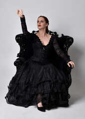 full length portrait of  woman wearing black gothic dress, sitting on a ornate black armchair. Seated pose, against a studio background.