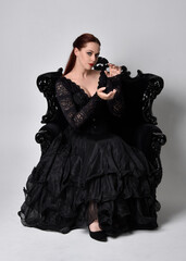 full length portrait of  woman wearing black gothic dress, sitting on a ornate black armchair. Seated pose, against a studio background.
