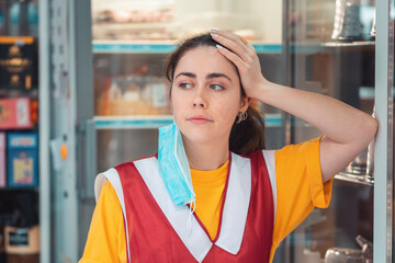 Portrait of a bored employee in uniform, with a medical mask over his ear. Showcase with products in the background. Concept of economic crisis during the coronavirus pandemic