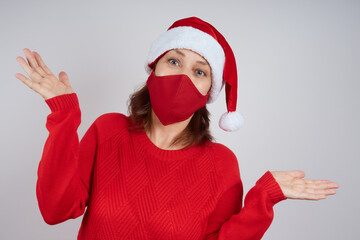 Fototapeta na wymiar An adult woman in a red protective mask, in a Santa hat on a gray background, spread her arms in surprise. Christmas, New Year in quarantine