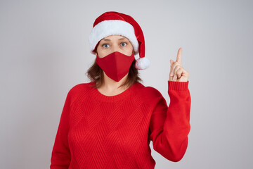 Fototapeta na wymiar A mature woman in a red protective mask, in a Santa hat on a gray background, shows a gesture of attention. Christmas, New Year in quarantine