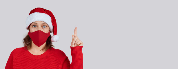 A mature woman in a red protective mask, in a Santa hat on a gray background, shows a gesture of attention. Banner with place for text, copy space