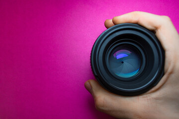 Holding camera lens in hand with blue reflection and colorful neon glares against purple background with space for text or logo. Top view
