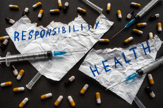 Danger of drugs to life concept. Crumpled burnt sheets of paper with inscription "Health" and "Responsibility" on black background with used syringes and many pills