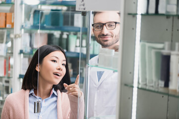 curious asian customer pointing with finger near bearded pharmacist in drugstore