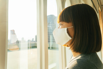 Side view - Beautiful young asian thai black short hair woman wearing white surgical mask looking outside the window.