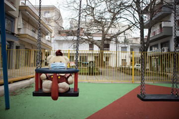 Close up shot to a teddy bear. It wears a blue mask at an empty playground. On a baby swing. Protection against coronavirus disease (COVID-19) outbreak.