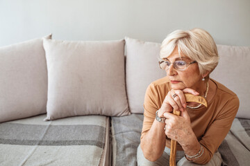 Senior woman holding a walking stick. Elder lady sitting on the couch with wooden walking stick. Retired woman with her wooden walking stick at home