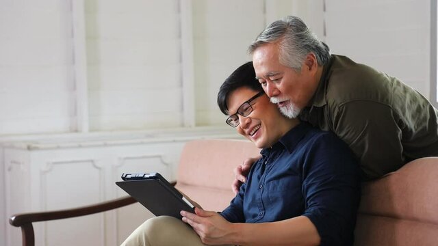 Asian senior man learn how to use digital tablet from his son while sitting on the sofa couch at retirement home