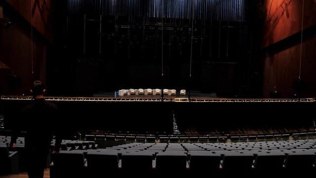 Rear view of a man walking down the stairs inside the empty dark hall for a conference or a forum. Media. Empty concert hall before mass public event with a lot of empty chairs and comfortable beige