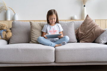Little girl sitting on the sofa playing digital tablet in the living room at home