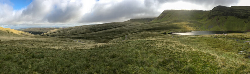 A panoramic view of the rolling vivid green hills and mountains of the Brecon Beacons in Wales with a lake in the foreground.