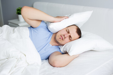 portrait of tired young man lying in bed and holding pillows on head