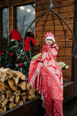 Fototapeta premium The girl is wrapped in a red blanket and is sitting on a hanging swing. New Year's decorations.