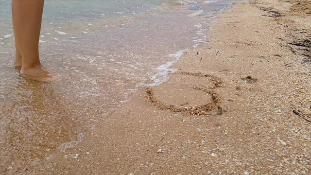 The wave washes away the heart nestled in the sand. Woman draws a heart in the sand. A woman draws a heart on the seashore which is washed away by a wave.