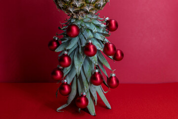 Original Christmas tree concept. Red shiny glass baubles hangs on the leaves of ripe pineapple standing upside down. Red festive background.