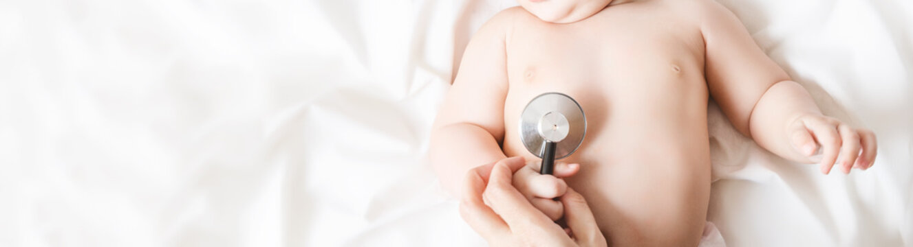 Pediatrician Examining A Toddler With A Stethoscope. Little Baby Lies On A White Bed. Health Concept. Child Holding A Stethoscope In A Small Hand. No Face Visible