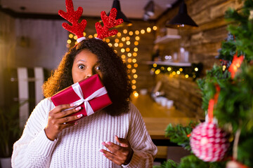brazil brunette hair woman hold red gift box near fir-tree at her apartment she is wear white sweater and red deer horns