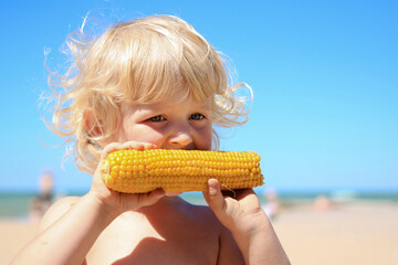 Little blond boy eating boiled corn on the beach. Healthy food, summertime, vacation concept.