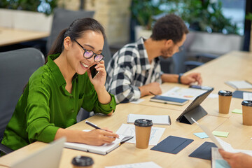 Young cheerful asian woman talking by phone and making some notes while working in the modern office