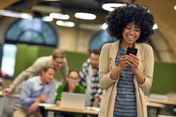 Young happy mixed race female office worker looking at smartphone and feeling excited while standing in coworking space, colleagues on the background