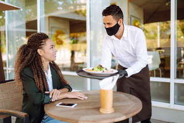 Young waiter in protective face mask and gloves with ordered meals, ready to serving guest. Waiter in cafe during coronavirus outbreak. Covid-2019.