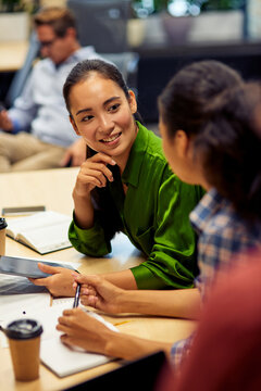 Vertical Shot Of A Young Cheerful Female Office Worker Discussing Something With Her Colleague While Sitting At The Desk And Working Together In Coworking Space Or Office