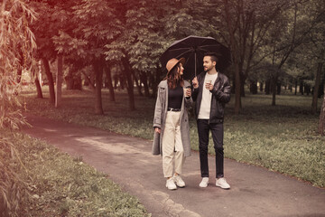 Fototapeta premium Young couple walking in the spring green city park, drinking hot coffee paper eco recycle cup. Two lovers husband and wife spending time together, discuss news or film movie with black umbrella.