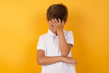 Cute Caucasian little boy standing against yellow background  making facepalm gesture while smiling...