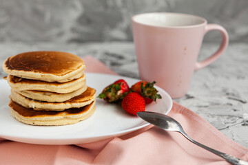 A stack of pancakes with a pink tea mug and fresh strawberries on a white plate on a light background. Horizontal orientation. Homemade american pancakes. Copy space.