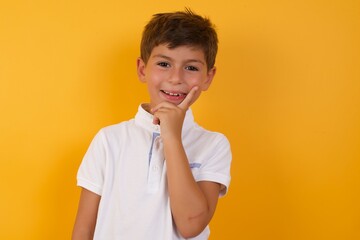Carefree successful Young handsome Caucasian little boy standing against yellow wall touching jawline gazing camera tilting head grinning white teeth delighted. Dental care concept.