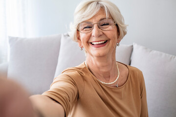 Selfie Portrait of Happy Senior Woman With Gray Hair at home. Portrait of elderly woman with short white hair wearing white glasses and light blouse holding camera and taking selfie.