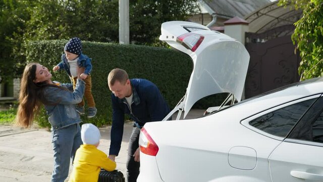 family car trip, joyful parents with their male children put suitcases in trunk for picnic out of town on weekend