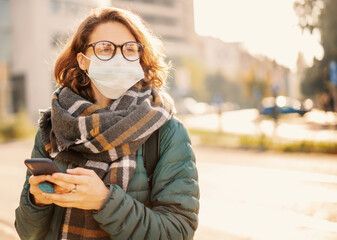Portrait of a beautiful young European woman in a protective mask on a city street with a smartphone in her hands. Antiviral measures in a pandemic, health and safety new normal life