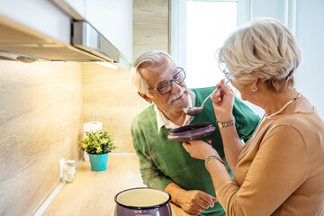Senior couple enjoying while preparing lunch together in the kitchen. Retired people at home preparing lunch with recipe book about bio vegetables. Happy elderly concept with mature pensioner
