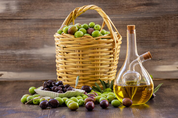 Green olives in a basket on a wooden floor, black and green olives on the ground, olive oil in a bottle, black olives in a bowl.