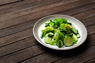 Closeup of plate full of green fresh vegetables, salad dish.Pieces and slices of cucumber,marrow, sweet pepper, broccoli salad on the plate,wooden table.Empty space