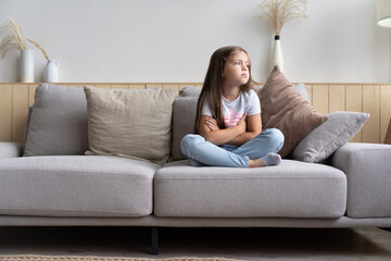 Beautiful unhappy sad little 6-year old girl sitting cross legged on couch in living room, feeling bored and lonely, being home alone while her parents spending all day at work