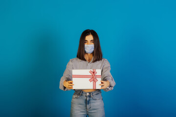 Fototapeta premium Young woman wearing protective medical mask holds white gift box with red bow isolated on blue background. New year gifts during coronavirus.