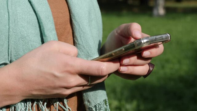 A woman in the park is surfing through the news feed on her phone, scrolling through photos, enlarging the picture. Hands close-up.