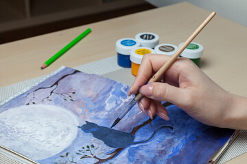 Female hands hold a brush and a cover from a watercolor jar and mix paints against the background of a cat on a tree, the night sky and moon near work tools on the table. Fine art in quarantine mode.
