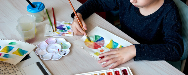 Boy painting pictures with watercolor paints during art lesson. Pupil on drawing with brush. Watercolor color wheel and palette. Color theory beginner hobby lessons.
