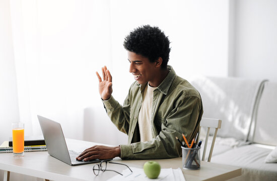 Distance communication concept. Positive black teen guy talking to his tutor or fellow students, waving at laptop webcam